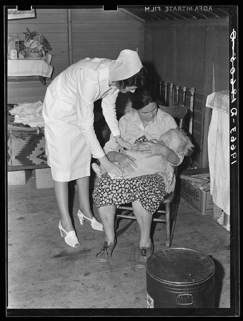 The image depicts a scene from the past, where an elderly woman is seated on a chair with a young child resting in her lap. She appears to be attending to the needs of the baby while another older individual stands beside them. This moment seems captured inside a makeshift medical tent or shelter made out of various materials such as wooden panels and fabric coverings.

The atmosphere evokes feelings of tenderness, care, and support among these individuals who are possibly family members or close acquaintances providing aid to one another during challenging times. The overall setting suggests that this might be an emergency situation where the group has gathered together for assistance related to health concerns involving children within their community.