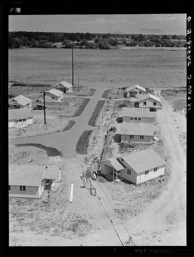 The image is a black-and-white aerial photograph of a rural area, showcasing several small one-story houses with pitched roofs. These dwellings are arranged in an irregular pattern on what appears to be a dirt road or path that cuts through the community.

In between and around these homes, there are some larger structures such as barns or workshops. The landscape is flat with sparse vegetation, indicative of farmland or agricultural land use nearby. 

There's a mix of utility poles connected by wires, suggesting electricity infrastructure for the residences. A truck is parked in front of one house on the left side.

The photograph provides context that these houses were built as temporary shelters for agricultural workers to enable them to settle permanently close to their places of employment in agriculture. The cost mentioned is eight dollars and twenty cents per month, including water and electricity. This tract lies adjacent to a migratory labor camp located at Farmersville, California.