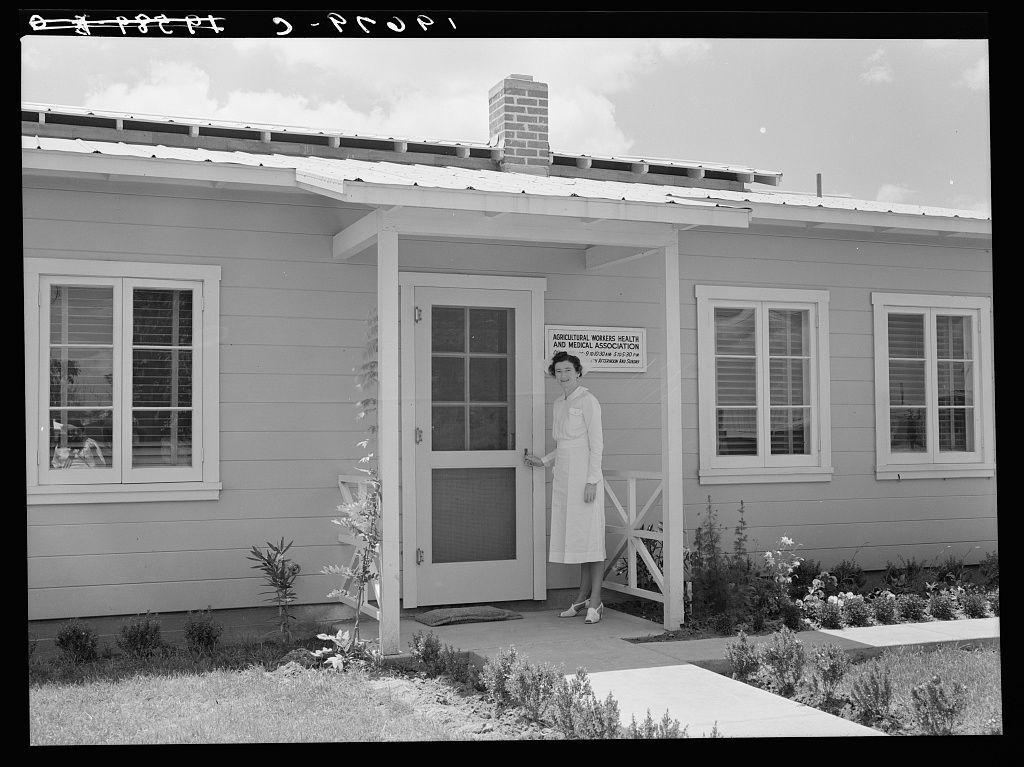 A black-and-white photograph depicting a woman dressed in white nurse attire standing outside of an old-style building labeled as the Agricultural Workers Health and Medical Association. The structure is simple, with multiple windows on its sides and front-facing door that has both glass panes and metal grills. Surrounding flora appears to be well-maintained hedges and flowering plants suggesting a cared-for environment. The photograph captures this scene during daytime under clear skies as indicated by the brightness in the image.