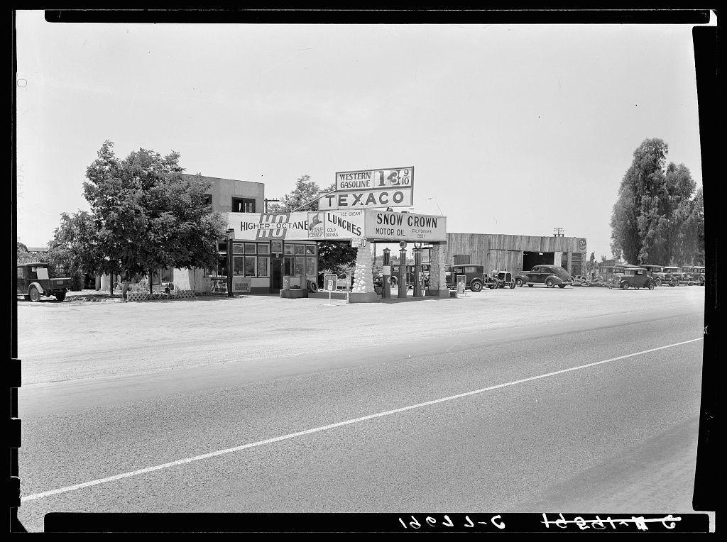 The image depicts a vintage black and white photograph of an old gas station on the side of a road. The Texaco service station is prominently featured in the center, with signs displaying gasoline prices at 16 cents per gallon for Western grade fuel and higher octane H.O. gas available as well. There are additional signage indicating Snow Crown motor oil from Standard Oil Company (Continental) USA.

The photograph appears to be taken during a time when cars were more abundant on the roads, likely in the early-to-mid 20th century given its style and appearance. The Texaco station is surrounded by an open area with sparse vegetation; there are trees visible but no buildings or significant infrastructure other than the gas pumps.

The road stretches out into the distance, hinting at a rural setting as it seems to be quite empty of traffic beyond this point. A few cars can be seen parked in front of the station and along the side of the roadway, suggesting that they belong to customers who are inside purchasing fuel or other goods from what appears to be a small shop attached to the gas pumps.

There is also visible text at the bottom right corner with "10843-6 Hedrix 2" possibly indicating some form of cataloguing system for photographic negatives. The image has been labeled as belonging near Kingsburg, which suggests that this location could have historical or geographical  [...]
