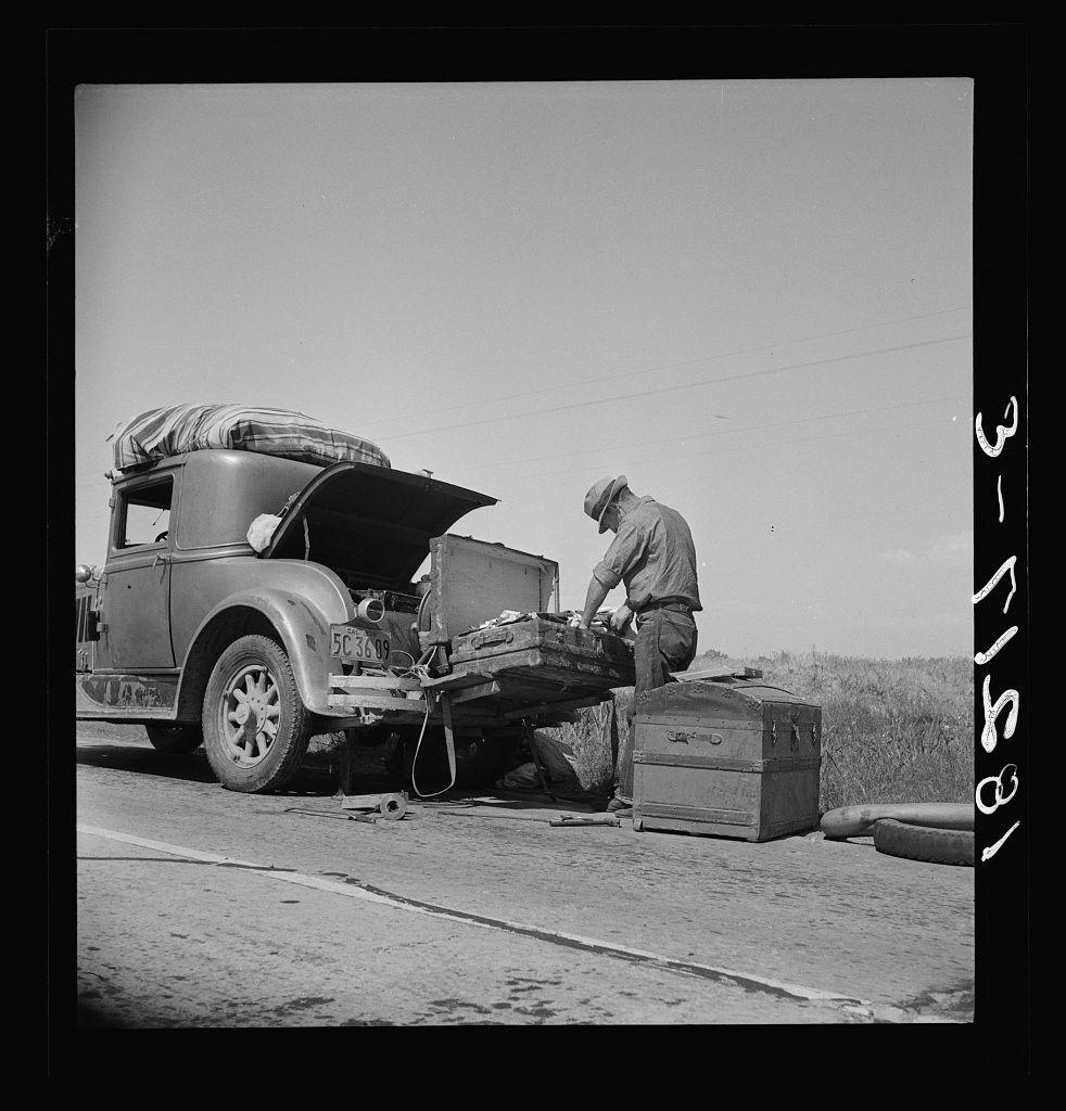 The image is a black and white photograph depicting an older man working on the side of a road. He's attending to what appears to be mechanical issues with his vintage car, which has its hood open revealing the engine compartment. The vehicle seems old-fashioned, likely from early mid-20th-century design given by visible features like round headlights and simple bodywork.

The setting is rural or semi-rural as indicated by a barren field in the background under clear skies suggesting it could be hot due to lack of cloud cover. On top of the car's luggage rack rests what seems to be bedding, indicating either travel or preparedness for an overnight stay.

Next to him on the ground are two large wooden boxes that he is working with; one appears opened as if its contents have been removed and laid out in front of it while his hands engage a tool. His posture suggests concentration and effort towards fixing what has presumably gone wrong with his car's engine or some other mechanical part.

The overall atmosphere conveys hardship, manual labor, resilience against adversity, potentially economic struggle suggested by the text on the image which indicates this may be a depiction from rural life during tough times like Great Depression era in California. The presence of luggage suggests that he might be on long travel journeys possibly related to work or migration.

Textual deta [...]