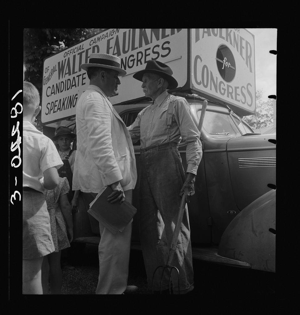 This black-and-white photograph depicts two men engaged in a conversation on the side of an outdoor event. One man is dressed formally, wearing a hat and suit with his hands clasped together while holding what appears to be a book or pamphlet. The other man stands next to him, clad in more casual work attire including overalls and boots. Behind them are large signs supporting political campaigns for General Walter Faulkner as candidate speaking, along with another sign indicating support for "Haulkner" running for Congress. In the foreground of this scene is a young child who seems curious or interested in what's happening around him. The presence of multiple onlookers suggests that some form of public event is taking place during which these interactions are occurring.