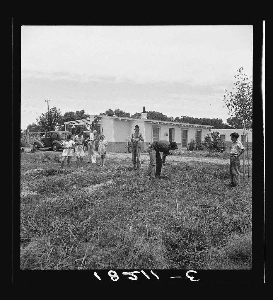 The image depicts a group of people, including both adults and children, gathered in an outdoor setting with sparse vegetation. The scene appears to be from the 1940s or early 1950s based on their clothing style, which includes dresses for women and suits for men.

In the foreground, two individuals are bending over as if engaged in gardening activities; one person is using a tool, possibly tilling soil. Behind them stand three people observing, including two adults with children beside them—two girls wearing similar white outfits, suggesting they might be sisters or close friends. Another adult stands slightly apart from this group.

In the background, there are several structures: a single-story building that resembles a farmhouse and another smaller structure to its right. A vintage truck is parked near these buildings, indicating transportation of goods or people between locations.

The landscape appears rural with an open field in front of the occupants' residence. The sky above seems overcast, which adds to the overall somber atmosphere of the image. Notable features include a telephone pole and lines stretching across parts of the scene from left to right, suggesting communication infrastructure for the remote location where this photo was taken.

A date is visible on the bottom edge of the picture in Roman numerals: "XVIII 1942," which indicates when the photograp [...]