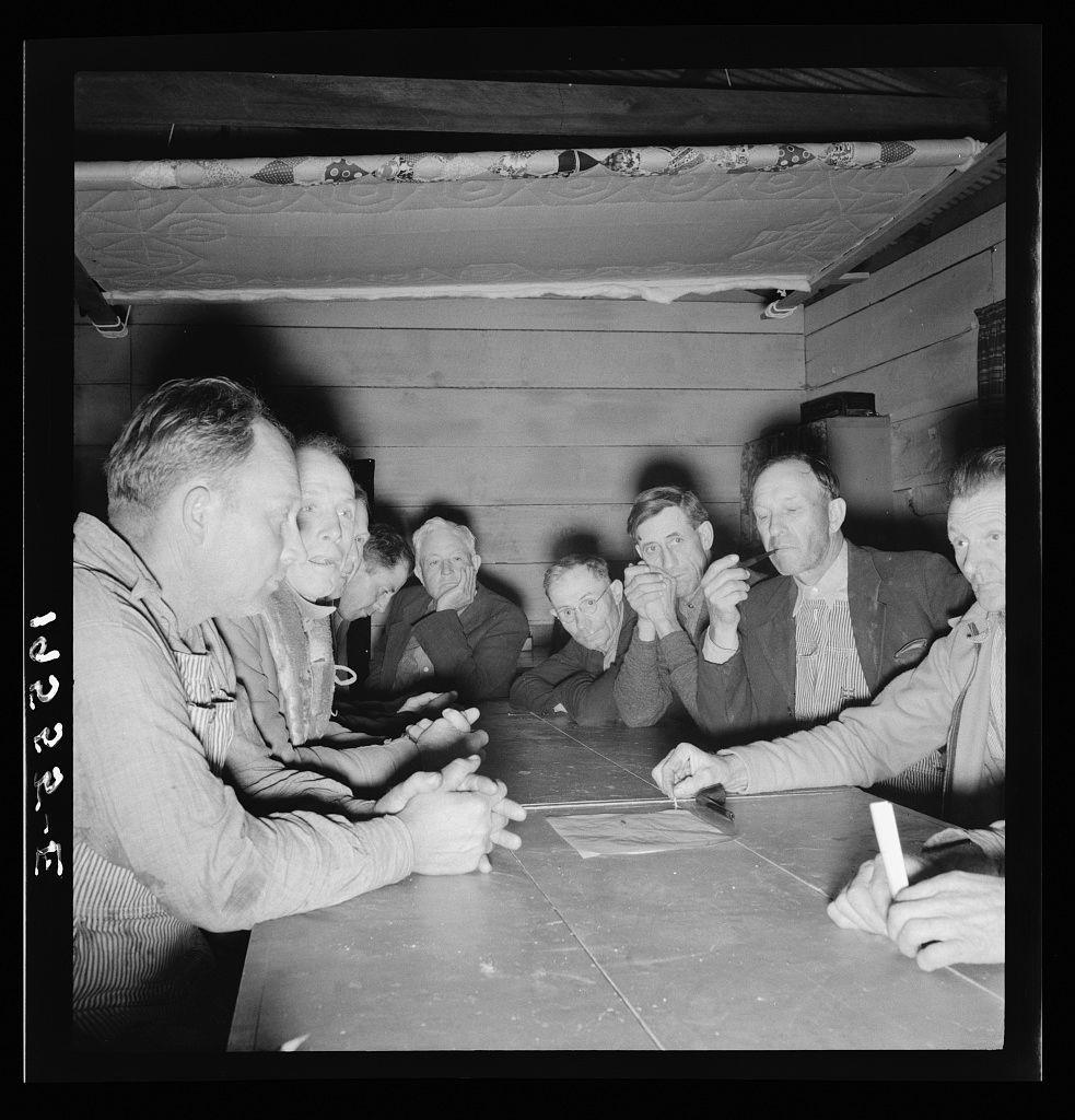 The image depicts a group of seven men seated around a table engaged in what appears to be an intense discussion or meeting. The setting seems informal, possibly within the confines of a farm building given its rustic wooden interior and presence of quilts on the wall above them.

Each man is dressed differently but generally casually; some wear overalls while others sport jackets with varied patterns. They are focused on something in front of them which could be papers or notes spread out on the table, suggesting they might be discussing work-related matters such as production quotas, financial issues, or community concerns common to a farm labor setting.

The photograph is black and white, indicating it's likely from an earlier era when color photography was not widely used. The monochromatic tone lends itself well to highlighting contrasts between subjects' faces and clothing against the stark interior background of what could be considered drab by modern standards but which would have been typical for rural farm life during certain historical periods.

The presence of writing tools in their hands suggests active engagement, perhaps taking notes or making decisions. Their expressions range from thoughtful contemplation to serious concern; this mix reflects a community dealing with challenges and striving towards solutions together. 

Overall, the photograph captures a  [...]