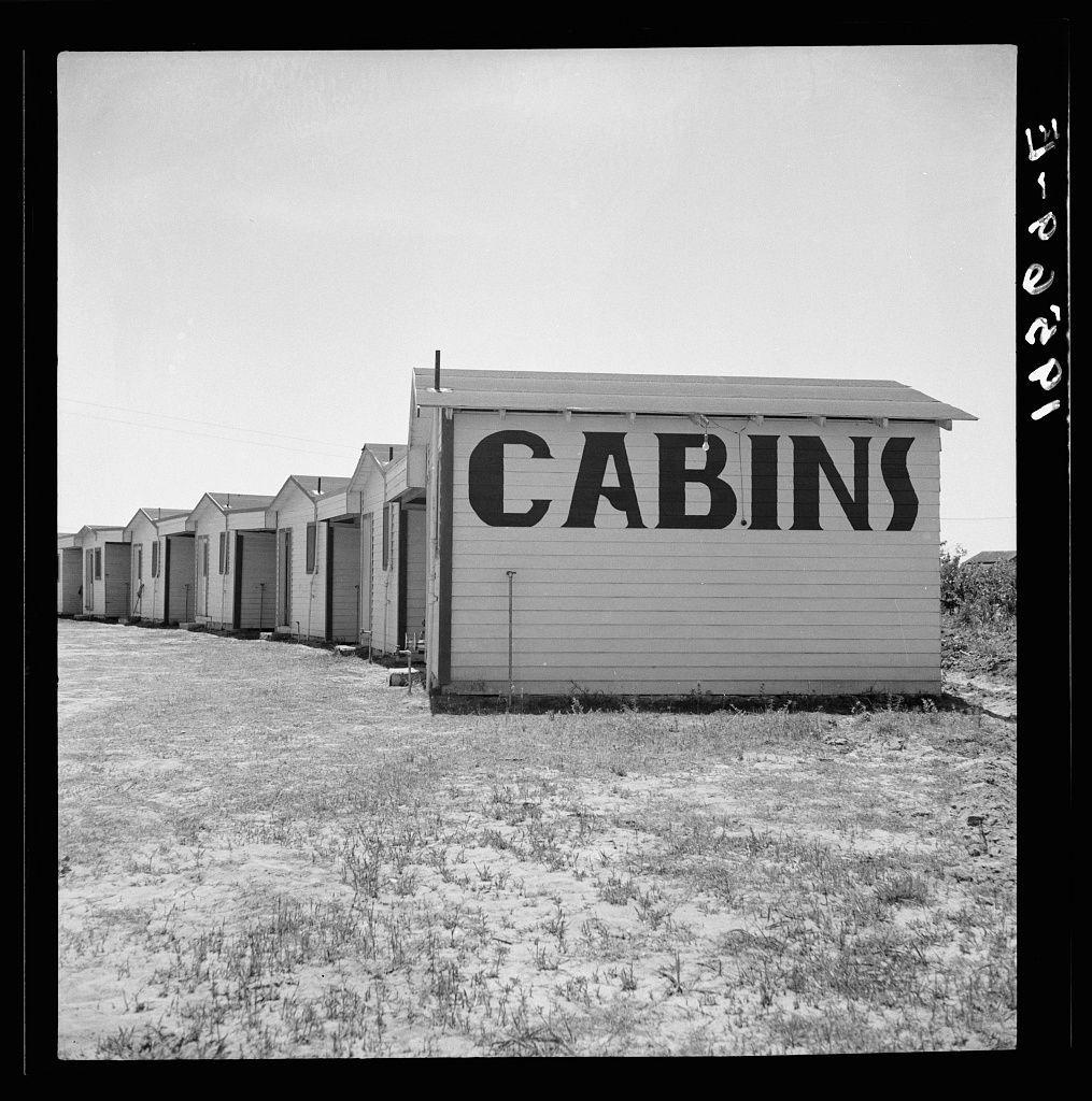 This black and white photograph displays a series of small, single-story cabins aligned in a straight line on what appears to be an open field. The ground is barren with sparse vegetation indicative of arid or drought conditions. Each cabin has a simple facade featuring two windows—one near the top and one closer to the middle—and a closed door at its center. Prominently positioned among these structures, facing towards the camera, stands another building larger than the rest, bearing large block letters spelling "CABINS" in bold typeface on its side.

The sky is overcast with no visible clouds, casting an even light across the scene and enhancing the high-contrast nature of a monochrome photograph. The lack of human presence or activity suggests abandonment or disuse. Given that it's black and white photography without color cues for time period identification, specific details regarding age are difficult to ascertain.

The setting is devoid of any other notable landmarks or structures beyond these cabins, which reinforces the sense of isolation in a vast open landscape. The image captures an essence of desolation and solitude amidst what appears to be a temporary lodging setup along a route not currently bustling with activity.
