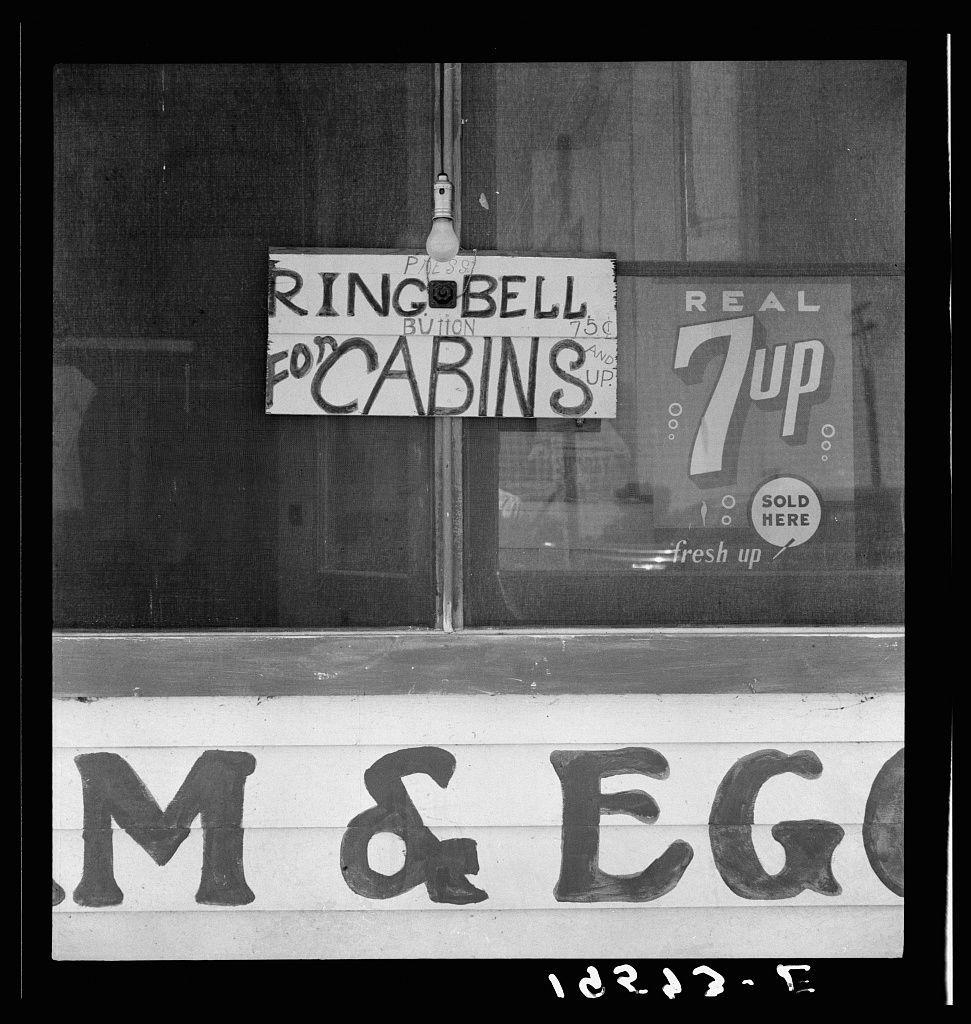 The image is a black and white photograph depicting an urban storefront, possibly from the mid-20th century given its style. The window features two signs with handwritten lettering: one instructs to ring for cabins on Bell button, which suggests it's likely at or near such accommodations; another advertises Real 7up soda as freshly sold up.

Below these signs is a larger painted sign reading "M & E" in bold capital letters, possibly indicating the name of the establishment. The backdrop includes what appears to be part of an electric bulb and various other indistinct items reflecting off or within the window.

The photograph's grainy texture suggests it might have been taken with film photography equipment common during that era. There is also a sequence of numbers at the bottom, potentially referencing documentation related to this image in archival context. The overall setting seems nostalgic, offering insight into commercial practices and visual design from its time period.