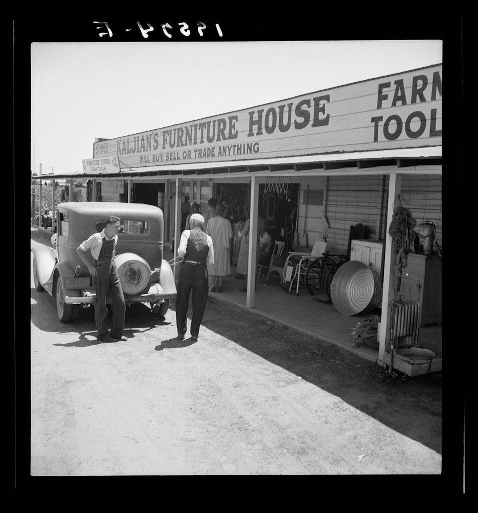 Black-and-white photograph depicting a storefront labeled "Kaljian's Furniture House" and "FARm ToOl". Two men are standing beside an old-fashioned vehicle, with one seemingly inspecting the tire. Inside the store, various items including chairs and wheelbarrows can be seen displayed on shelves. The setting appears to be in California during early-to-mid 20th century based on architectural style and attire of individuals present.