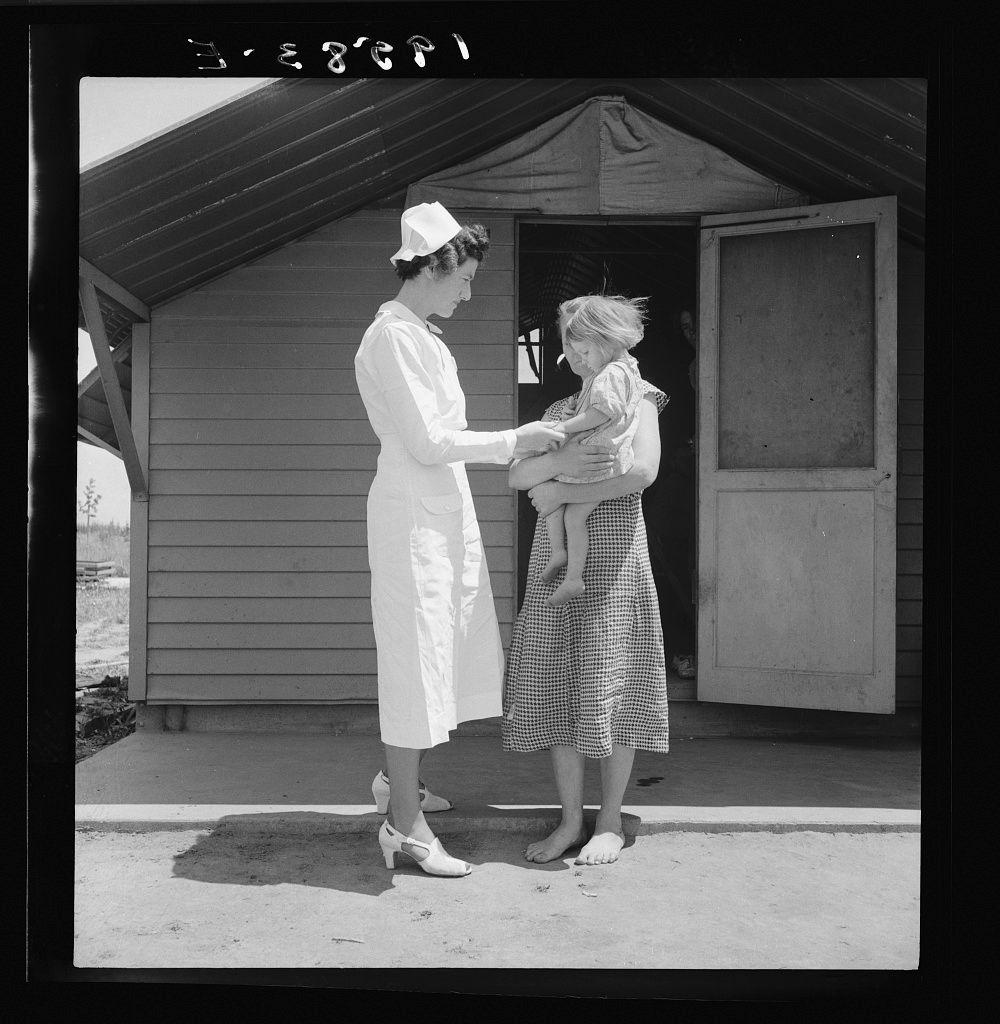 The image depicts two women and a child in an outdoor setting. On the left, there is a woman dressed as a nurse wearing a white medical uniform with a cap, holding what appears to be a bottle or small container towards another woman on her right side who stands beside a house-like structure. The second woman wears a knee-length dress adorned with black and white checks and holds a young child in her arms; the child is wrapped in a blanket. Both women look down at an unseen point, possibly engaging in conversation about the baby's health or care. In the background, we see part of another building which seems to be connected by a wooden structure, suggesting this scene may take place on someone’s property rather than within a clinical setting. The image is black and white with clear contrasts that enhance its historical feel, indicative perhaps of mid-20th-century photography.