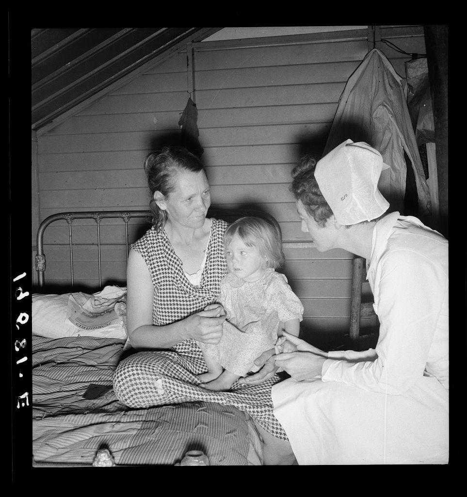 The image is a black-and-white photograph depicting three individuals in an indoor setting, likely from the early to mid-20th century based on their clothing and hair styles. Two women are sitting with one child between them; both women wear patterned dresses or nightgowns, indicating they might be at home during leisure time. The woman holding a young girl is smiling gently while looking down towards her hand resting in the other's lap. Beside them sits another older woman wearing what appears to be a nurse’s uniform with an apron and cap, engaged attentively as she examines or assists the child.

The background shows part of wooden paneling on one wall and a partially visible bedframe where clothing items can be seen laid out. The overall setting suggests a domestic scene possibly involving health care within a home environment for rural residents in Farmersville, California during FSA's camp period mentioned at the source link provided below it.