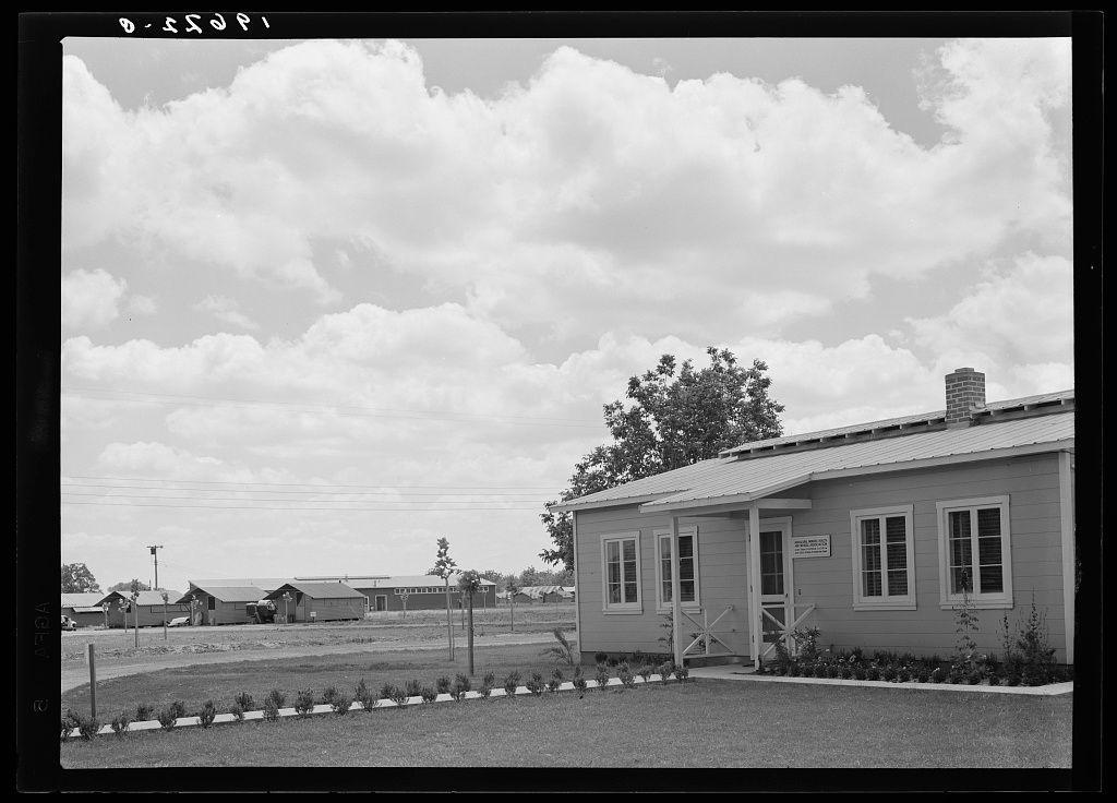 The image is a black and white photograph depicting an outdoor scene with significant cloud coverage in the sky. In the foreground, there's a modest one-story building that appears to be made of wood or metal siding. It has a simple rectangular shape with two visible windows on its side facing the camera; each window features shutters but no apparent curtains or blinds. The front entrance is sheltered under an extended eave supported by wooden posts and includes what looks like a small porch area, suggesting that this building might serve as a reception hall or entry point to another structure.

Adjacent to the main building are several smaller structures with gabled roofs; their use isn't immediately clear but they could be storage sheds or residential units. The ground is partially covered by grass, and there's a neat row of white posts along one side that seems like an informal fence dividing different sections within this area. In front of these buildings lies what appears to be paved grounds with some vegetation growing in patches.

The backdrop shows more structures at varying distances under the expansive sky filled primarily with fluffy clouds, indicating possibly fair weather conditions. There's a hint of greenery suggesting trees or bushes towards one end and part of an industrial structure is visible on the far right side of the photograph, which might indicate [...]