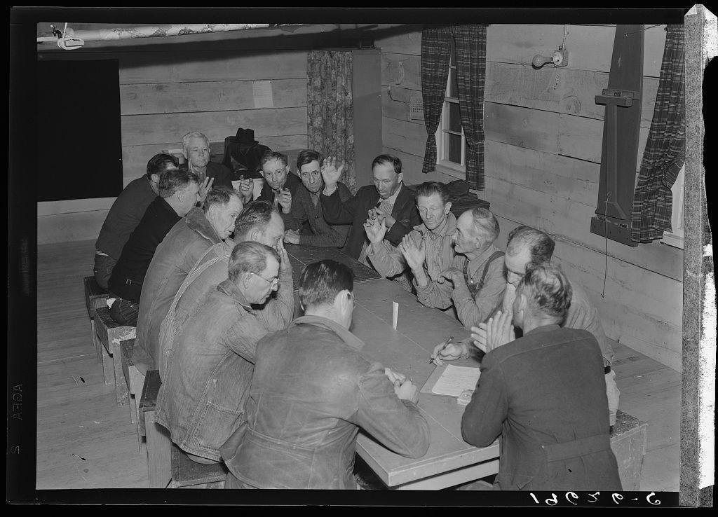 A black and white photograph depicting a group of people gathered around a table. They appear to be engaged in an intense discussion or meeting, with many participants raising their hands as if making decisions or voting on issues at hand. The setting seems informal, possibly within a camp for migratory agricultural workers, indicated by the attire of the individuals which includes work jackets and overalls typical of farm laborers from that era. Some are seated while others stand around the table, suggesting an open dialogue rather than a hierarchical structure.

The image is labeled with "Farm Security Administration (FSA) camp for migratory agricultural workers," followed by location details: Farmersville, California. The date stamp at the bottom right corner suggests it was taken in 1942 or later during World War II era when such camps were more common to house and manage migrant laborers.

The image captures a moment of collective decision-making or problem-solving within this community group, showcasing their active participation and engagement with one another.