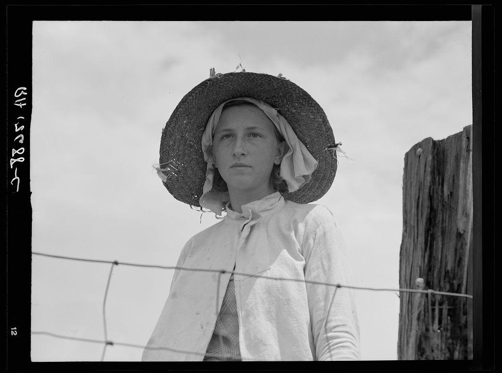 The black and white photograph shows a young woman standing against the sky, with her hands clasped behind her back. She is wearing an old-fashioned straw hat adorned with ribbons on either side of its brim, which are fluttering in what appears to be a gentle breeze. The same style of ribbon can also be seen tied under her chin and along the edge of her dress collar.

Her attire suggests that she might be dressed for rural work or travel: she wears what looks like a white cotton apron over an older-style blouse with puffed sleeves, which is tucked into her pants at the waist. The woman's expression is composed yet somewhat contemplative; there’s an air of pensiveness about her.

Behind her and to one side stands part of a wooden fence post, indicating that she might be on or near farmland property. In front of her, partially framing the shot in what looks like a barbed wire fence which suggests rural countryside setting. 

The photograph is labeled with handwritten text at its top left corner: "farmer's daughter in fields / farm Georgia" along with some numbers and letters that could possibly indicate metadata or categorization information related to this image within an archive.

This particular photo appears to be part of a larger collection, as indicated by the watermark which suggests it is from Farm Security Administration (FSA) photographs. The FSA was tasked during [...]