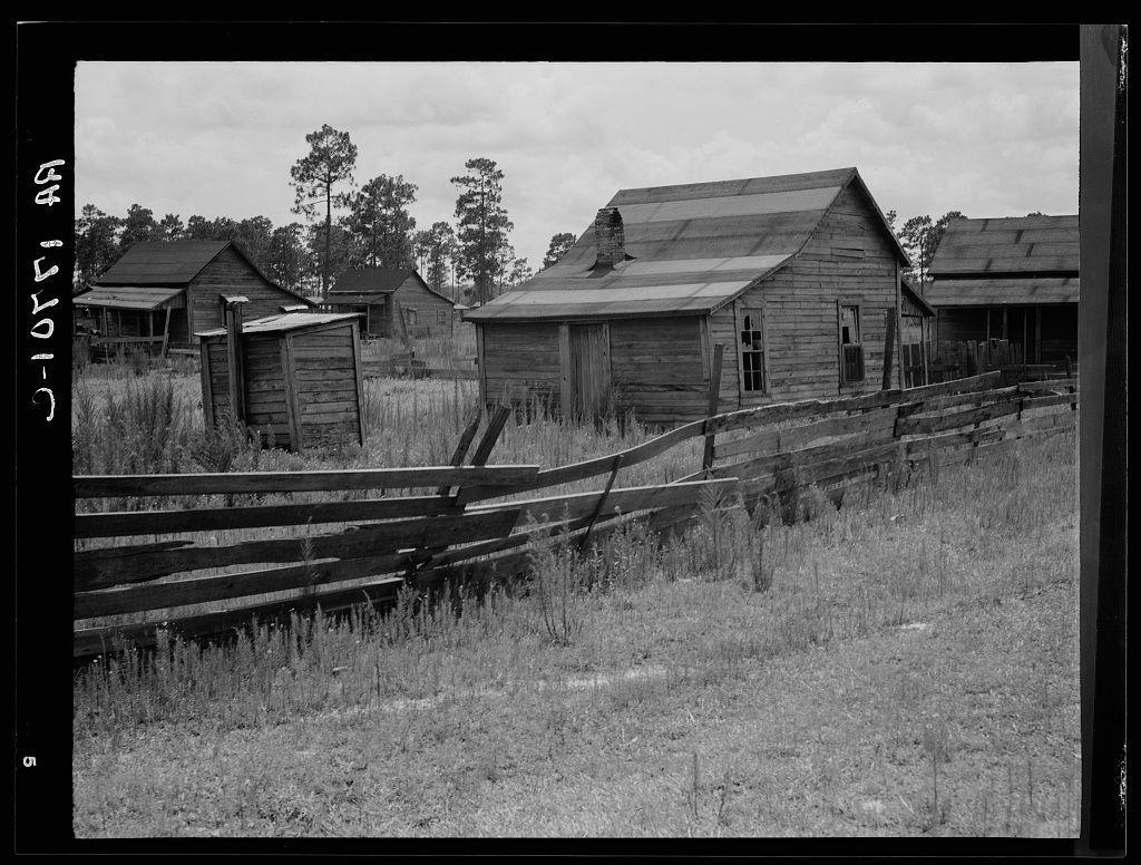 The image depicts a rural scene with an old, weathered wooden fence in the foreground. Beyond the fence lies a group of dilapidated structures that appear to be abandoned farm buildings or shacks. These buildings have slanted roofs and are constructed from planks of wood, some showing signs of decay such as missing parts and broken windows. The architectural style suggests these might date back several decades.

The landscape around is overgrown with tall grasses and weeds, indicating a lack of maintenance. There's an absence of modern infrastructure or vehicles, giving the scene a timeless quality. In the background stands a line of trees under what seems to be an overcast sky, contributing to the overall impression of abandonment.

In terms of geographical context, as mentioned in your additional information note about Careyville, northern Florida, this could potentially represent that area's rural past, possibly after the closure of local industries such as mills. This correlates with a historical period where agriculture was prominent and later saw shifts due to economic changes or natural disasters affecting farming communities.

The black-and-white format emphasizes textures and contrasts rather than colors, lending an air of nostalgia and emphasizing decay over vibrancy. It's important to note that while the image is described in detail here, it does not contain an [...]