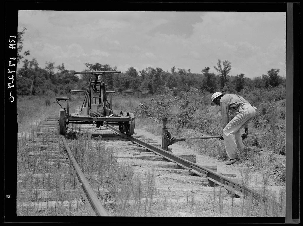 The image is a black and white photograph depicting an individual engaged in the task of removing railroad tracks from what appears to be a rural setting. The person, wearing a hat and light-colored clothing that suggests work attire possibly for protection against sun exposure or insects, bends over as they manipulate one of the rail pieces along the track's length.

The railroad equipment includes a manual lever mechanism mounted on wheels, likely used for loosening or lifting sections of the tracks. This machinery stands to the left side of the image and seems stationary in this scene.

Surrounding environment consists of what appears to be overgrown vegetation with no visible structures apart from some distant trees under an open sky, indicating a possible abandonment of industrial activity such as logging, given that it leads up to closed sawmill. There is also another set of railroad tracks parallel but not directly connected; however, they seem less maintained.

The photograph evokes themes related to laborious work in rural settings and possibly the transition from active industry (such as logging) leading to abandonment or deconstruction processes for removal of infrastructure like railroads.