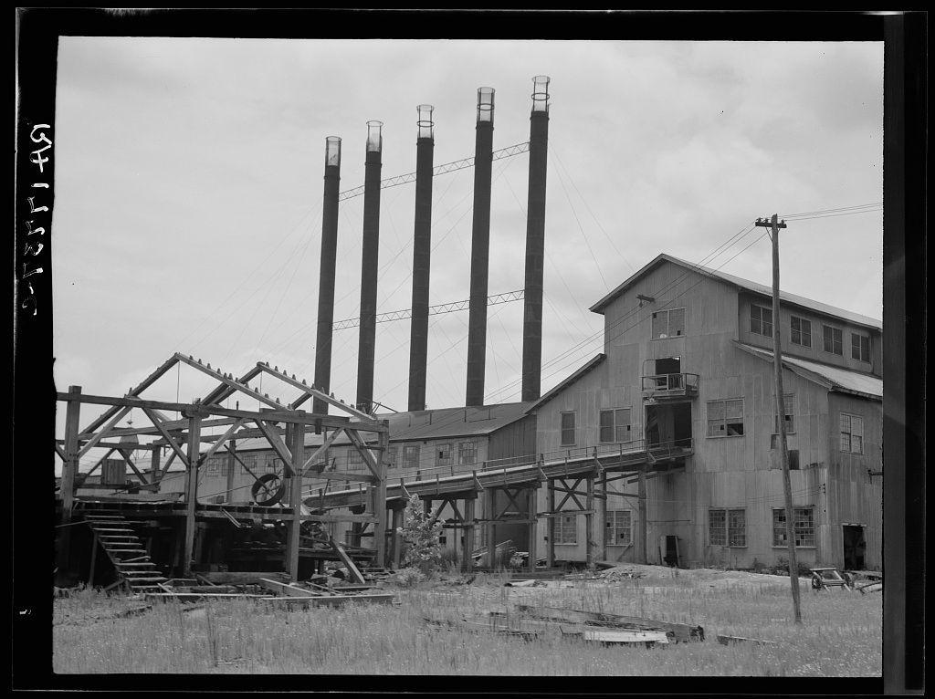 The image displays a black and white photograph of an industrial scene. In the center, there is a large wooden structure with metal beams that appears to be in disrepair or being dismantled, as evidenced by missing panels and exposed interior components. The framework includes several steel girders supported by columns, some without any decking, indicating areas for future construction work.

Surrounding this main structure are tall smokestacks made of dark-colored material which seem weathered over time. These stacks have a cylindrical shape with square bases at the top where they likely connected to chimneys or other ventilation systems inside the building complex. The sky is visible through gaps in what might be an incomplete roof, showing that it's daytime.

To the right stands a large two-story wooden building attached to another structure not fully captured within this frame. This industrial facility has numerous windows and appears aged with peeling paint on its exterior walls. There are electrical poles extending from behind the buildings, adding to the urban-industrial ambiance of the scene.

The ground is uneven with patches of grass or weeds growing through areas where materials might have been removed during dismantling activities. The overall setting suggests a site in transition between industrial use and potential future development or abandonment.
