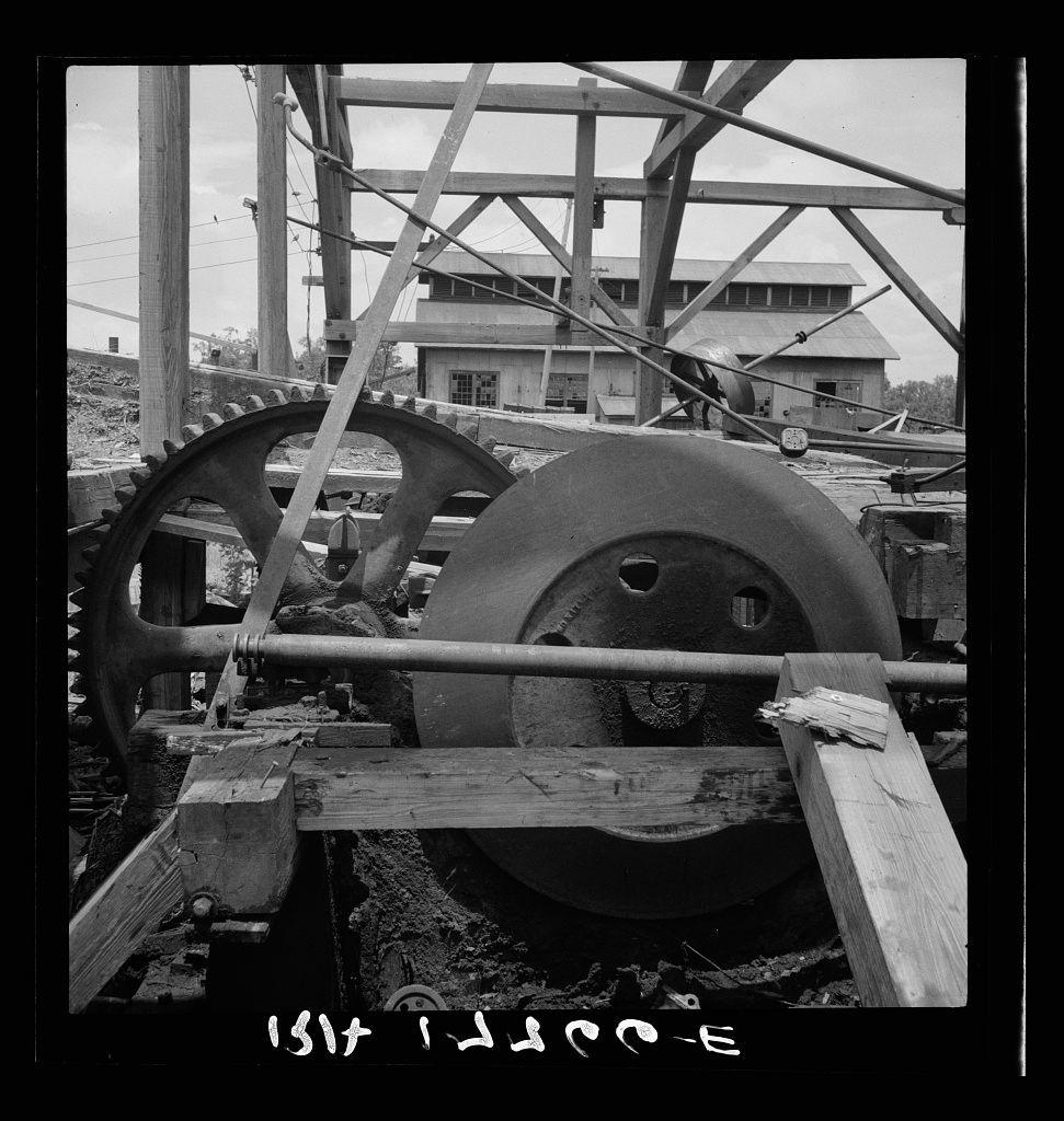 The image depicts a black and white photograph of an old-fashioned industrial machine that appears to be part of lumber processing equipment. This machinery includes large, interconnected gears made from metal with teeth on the edges for interlocking purposes.

In the background, there is some infrastructure possibly related to the operations; it's difficult to determine exactly what these structures are due to their somewhat blurred appearance in the photo. The setting seems to be outdoors as natural light and open sky can be seen above.

Various wooden beams or supports run across the scene, indicative of a dismantling process, which aligns with the caption indicating that this mill is being dismantled. There's visible dust and debris on some surfaces, suggesting active work in progress.

At the bottom left corner, there are handwritten notes: "1945" followed by what appears to be a series of letters or characters - it seems like an incomplete notation possibly relating to documentation related to this machinery. The photograph has a grainy texture typical of black and white film photography from that era.