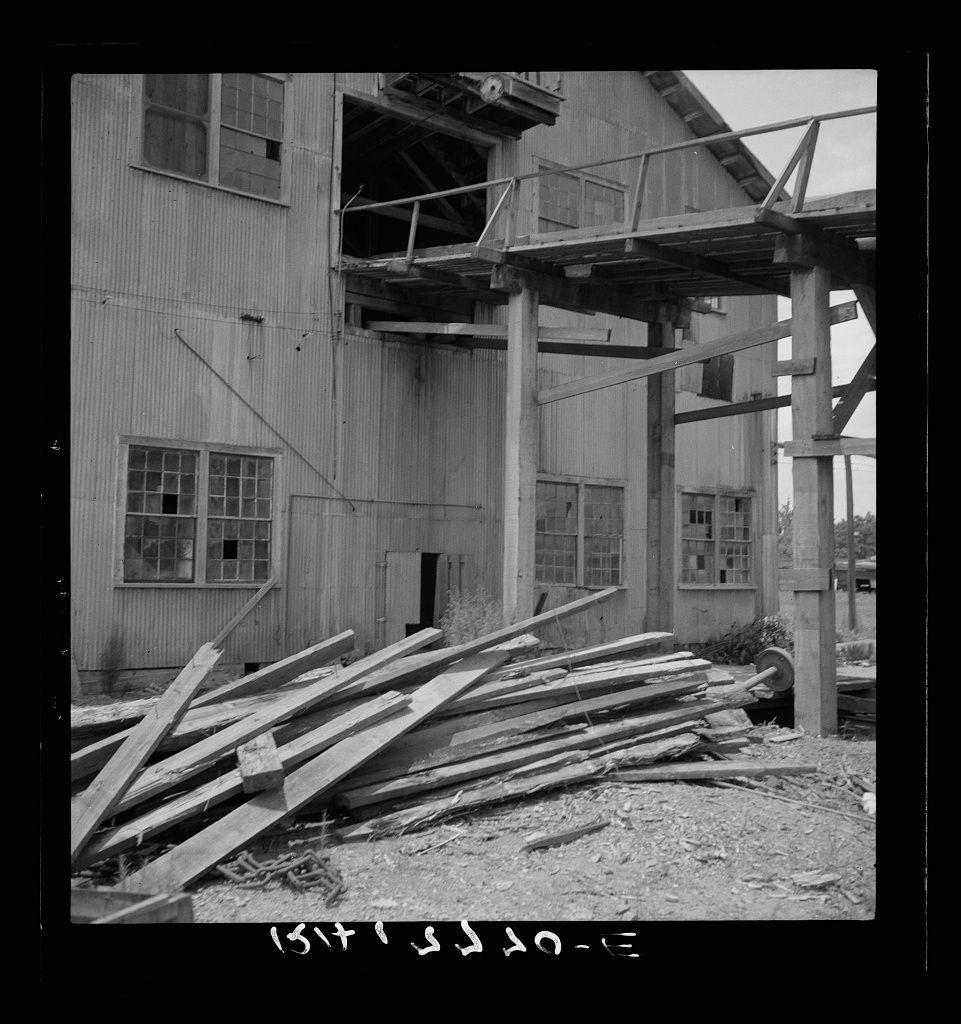 The image depicts a dilapidated industrial structure that appears to be in the process of being dismantled. The building is made primarily of corrugated metal, with various levels and exposed wooden beams suggesting it was once part of an operational facility, possibly related to lumber processing or manufacturing given its rustic appearance.

Several large pieces of timber are scattered around the ground outside the structure, indicating ongoing work on deconstructing it. A significant feature includes a wooden ramp-like extension that protrudes from one corner of the building, showing signs of wear and neglect.

The photograph is monochromatic, which adds to the sense of abandonment or decay. The sky above appears overcast, suggesting an atmosphere of desolation. There are no visible people in the image; it focuses entirely on the structure and its surroundings.

In terms of context provided by this single photo, one could infer that the building has been out of use for a considerable period, as evidenced by its deteriorating condition. The scattered timber implies recent or ongoing dismantling activities. Further information about this particular site can be found in relation to Careyville, Florida and is associated with Lumber mill structures being dismantled.