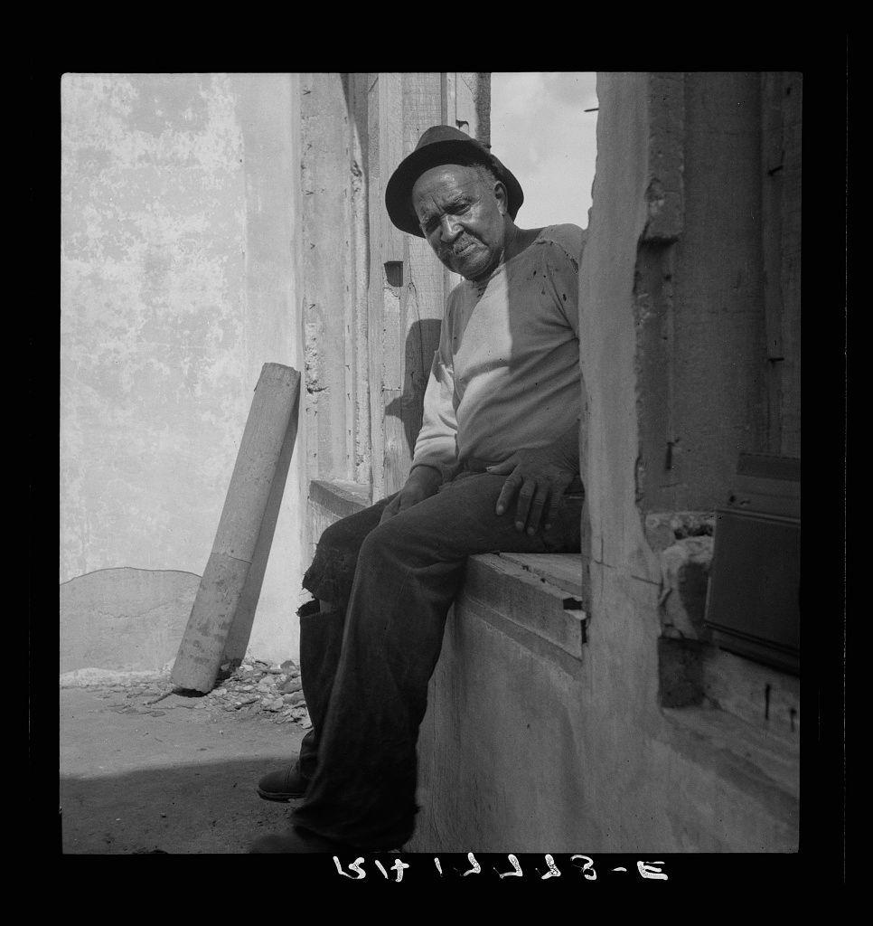 The image depicts a man sitting on the edge of what appears to be an old, dilapidated structure. He is dressed in casual clothing: a long-sleeved shirt with visible wear and tear at the elbows, dark pants, and shoes that appear worn out. The man's posture suggests he might be tired or reflective; his head rests against one hand while propping himself up on what seems to be part of a wall or platform.

The environment is stark and desolate, emphasizing abandonment rather than habitation. There are loose wooden planks and rubble around him, indicating neglect or decay over time. The photo's black-and-white composition adds to the somber mood, highlighting textures such as cracks in walls, peeling paint, and debris on the ground.

The image is labeled with a date "1938-5" at the bottom, which suggests it was taken during that period but could be an error or misinterpretation of historical data. The context provided by the caption reveals this photo to have been captured in Fullerton, Louisiana, once known as a lumber town and currently abandoned.

The overall mood conveyed is one of quiet endurance amidst desolation, likely evoking thoughts about the passage of time, abandonment, and resilience.