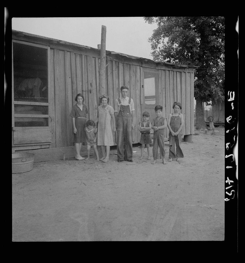 The image is a black and white photograph showing a family of nine standing outside on what appears to be sandy ground, with an older wooden building behind them. The structure has vertical planks and looks weathered, suggesting it may have been in use for some time. In front of the building, there's another smaller building or shed with its door open. Various household items such as a large pot are visible near this small shelter.

The family consists of two women on either side, one standing slightly behind an adult male figure who appears to be leading the group and is centrally located in the photograph. In front of these adults stand five children; three boys wearing overalls or shorts with button-up shirts, while two girls wear dresses that come down just above their knees.

All individuals are barefoot, suggesting a warm climate or lack of resources for shoes. The expressions on their faces range from neutral to slightly serious, and the overall posture is upright but not relaxed. It's possible they're posing for this photograph as part of a documentary project given its formal composition despite the seemingly humble surroundings.

The image captures what appears to be an ordinary moment in a rural setting with historical significance suggested by the context provided: it shows individuals affected by flooding, reflecting on their displacement and hardships during [...]