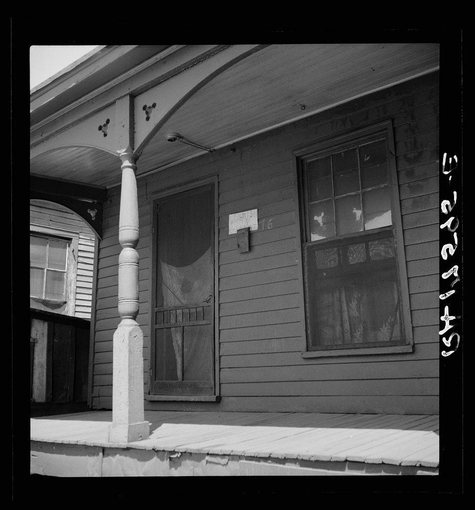 This black and white photograph shows a weathered wooden house with distinctive architectural features. The facade has two windows, one of which is partially obscured by curtains while the other appears to be covered in snow or frost. Between these windows, there's a small door marked '16', suggesting it could be an entrance way leading into a garage or storage space.

To the left, we see another section with boarded-up windows, hinting at neglect or abandonment. The porch is adorned by a robust stone pillar connected to decorative brackets and beams supporting the roof overhang above. These architectural elements suggest a vintage style possibly from the early 20th century.

The house's siding shows signs of aging; paint has worn away in several areas revealing wood beneath, and some sections are darkened or stained likely due to exposure to weather conditions. The door is slightly open with what appears to be curtains hanging inside it, yet no clear view into that room can be discerned from this angle.

There's a visible contrast between the house’s rustic exterior and its worn state against an unseen backdrop of urban decay. No people or modern elements are present in this scene; instead, the focus is on architectural detail and how time has affected residential structures.

No specific context about why it was taken exists within the image but given that "Furnish room [...]