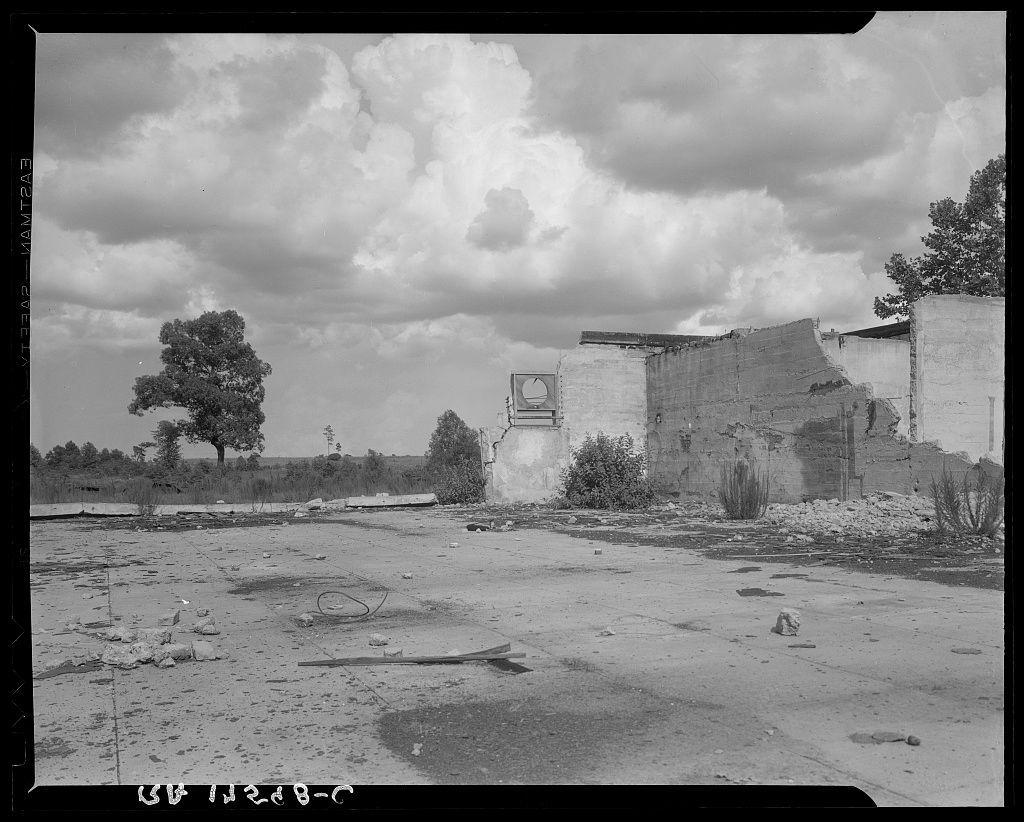 The image depicts an abandoned and dilapidated building in a desolate landscape. The structure appears to be partially collapsed, with large portions of the walls crumbling away. There's visible debris scattered around on what seems like cracked earth or concrete ground.

In the background, there is tall vegetation including trees, which suggests that this area might have been once inhabited but has since fallen into neglect. Above, a cloudy sky can be seen providing contrast to the stark and desolate scene below. The overall color palette of the photograph consists mostly of grayscale tones due to its black-and-white nature.

There's text on the top left corner of the photo which seems like an indication or location marker, but it is not entirely clear what specific details are intended by this notation.
