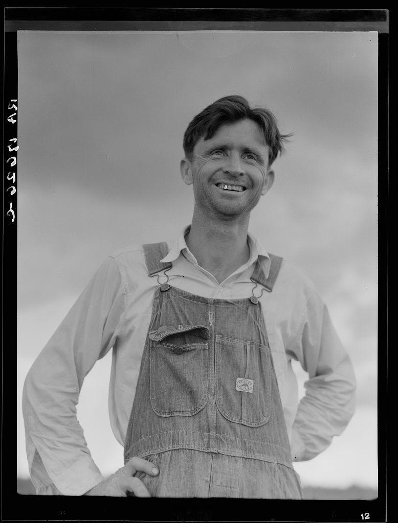The image depicts a man with a cheerful expression, wearing overalls and a light-colored shirt. His posture is relaxed, with one hand on his hip and the other resting against something out of frame. The setting appears to be outdoors under an overcast sky, which suggests that he might be in rural or agricultural surroundings given his attire.

The image seems historical, possibly from early 20th century America based on its black-and-white format and style. It evokes a sense of resilience and the human spirit during tough times, as suggested by the text mentioning the man's work history at Fullerton, Louisiana lumber mill for fifteen years before being left stranded in cut-over areas.

The photograph carries historical significance, potentially connected to social or economic issues faced during that period.