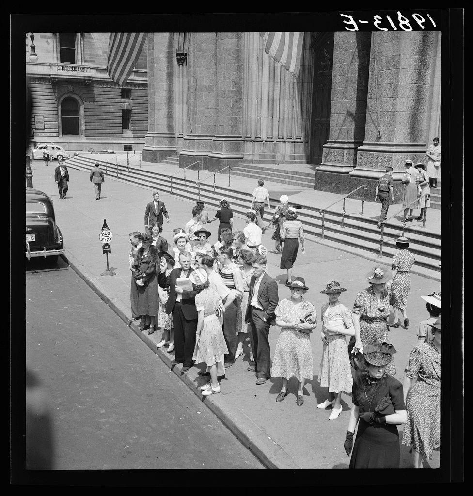 The image depicts a group of people, likely pedestrians or visitors, gathered on the sidewalk outside Saint Patrick's Cathedral in New York City. The black and white photograph suggests it was taken during an earlier time period, possibly mid-20th century.

In this scene, various individuals are standing together, seemingly waiting for something, such as an uptown bus mentioned in the provided caption or perhaps a scheduled event at the cathedral itself. Some of these people appear to be engaged with one another through conversation while others stand on their own, observing their surroundings.

The crowd is diverse, featuring both men and women dressed in attire that reflects early 20th-century fashion trends: hats for sun protection, knee-length dresses, and suits for some males. The presence of flags indicates a sense of patriotism or significance to the location they are visiting.

In addition to those standing on the sidewalk, there is also movement captured within the image as individuals walk by in different directions. A uniformed police officer walks past with his back turned towards the camera, adding an element of orderliness and safety to the scene.

The setting appears urban with well-kept streets and steps leading up to Saint Patrick's Cathedral's entrance. The architecture suggests a historical significance for this location within New York City, which is o [...]