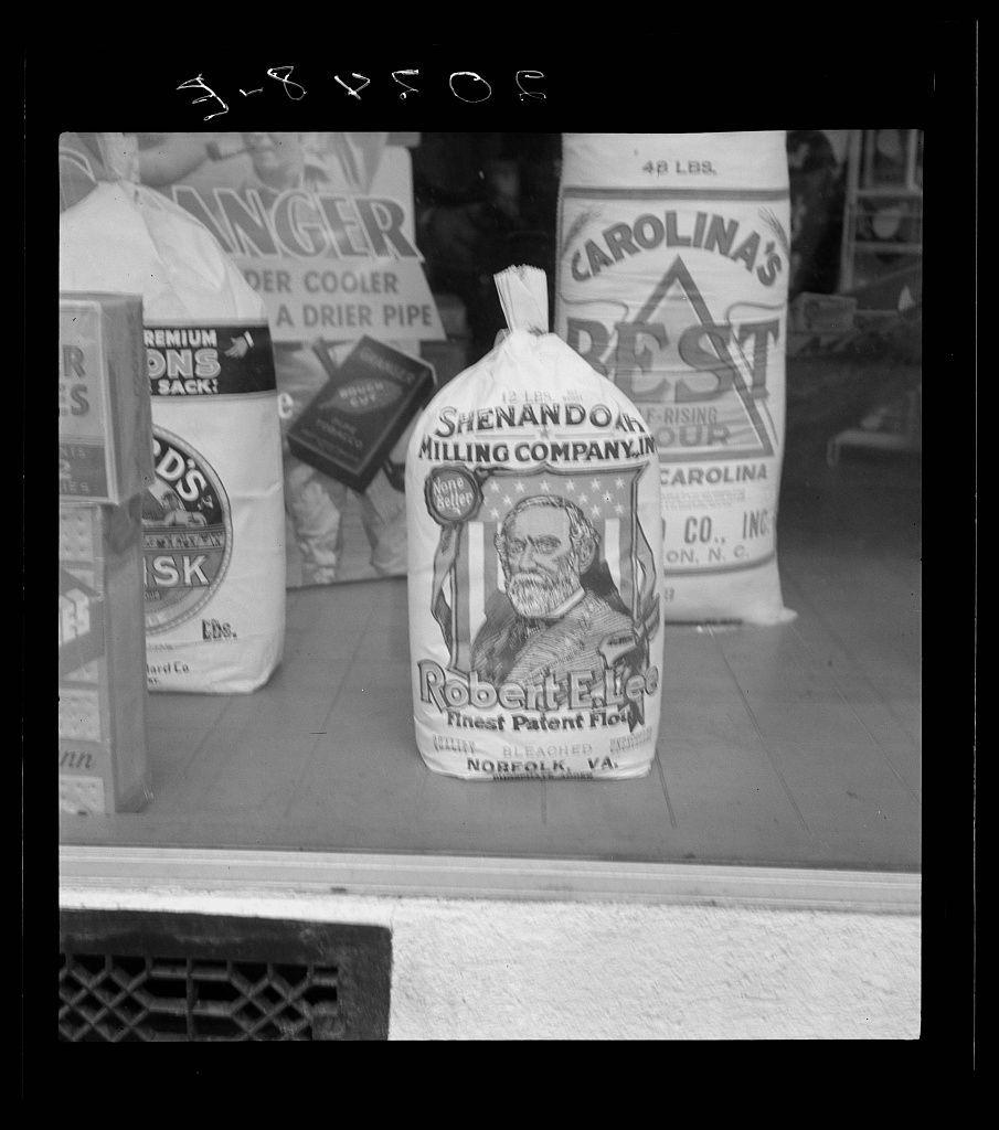 The image is a black and white photograph showing various bags of flour on display, likely from a grocery window. One prominent bag in the center features an illustration with text that reads "Rodgerson's Best Patent Flour," alongside additional details such as being "Finest Bleached" and originating from Norfolk, Virginia. The other visible products include large sacks labeled "Shenandoah Valley Milling Company Inc." which is a 48-pound bag of flour, and another branded "Carolina's", also indicating it weighs 48 pounds but the company name isn't fully legible due to perspective. There are various sizes and brands shown; some with price tags visible like "$1.05 LBS" on one sack, which suggests a retail setting where these goods were sold or displayed for sale. The background is mostly obscured by items in focus, but there appears to be a white vertical surface behind the products that could suggest store shelving or racks.