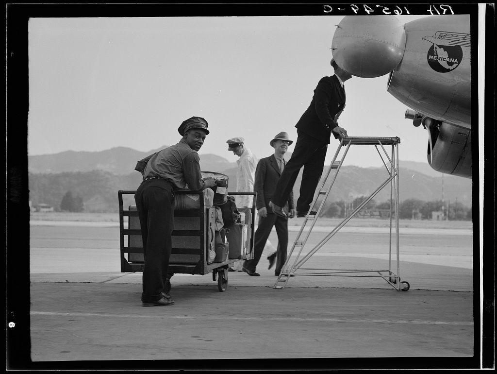 The image is a black and white photograph depicting four individuals interacting with an airplane on what appears to be an airport tarmac. From left to right, the first person is wearing dark trousers, a light-colored shirt, and a cap while pushing a luggage cart filled with various items of baggage. The second individual is dressed in formal attire, including a suit jacket, tie, dress pants, and shoes, standing on a metal boarding ladder that extends up to an airplane's door or window. To the right stands another person wearing a hat and coat, seemingly observing the scene, while behind them is someone with a light-colored cap and dark clothing, also partaking in this airport activity. In the background, we can see hills under a partly cloudy sky. The aircraft visible has branding that includes "Mexico-Air" and other unreadable text on its tail section.