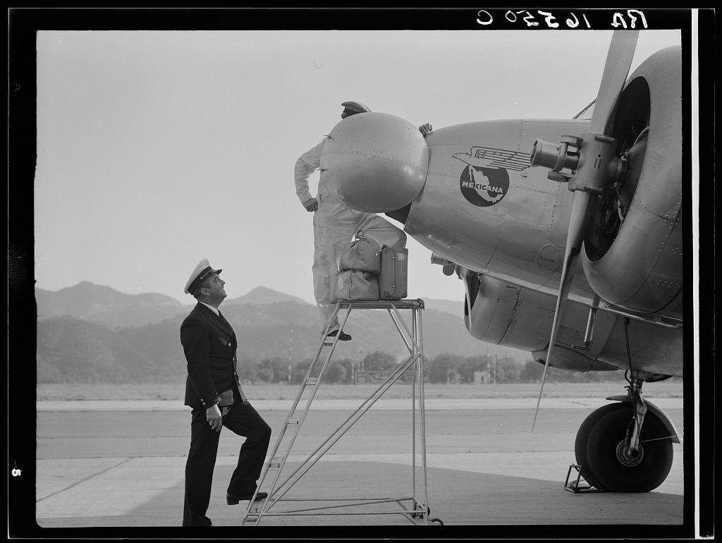 The picture depicts a moment on an airfield where two aviation professionals are engaged in what appears to be pre-flight preparations or inspection. One individual, dressed in formal pilot attire with a cap and dark suit, is standing upright, looking up towards the wing of an aircraft. He seems to be observing another person who is positioned higher than him, likely indicating that he's on a ladder platform extending into the plane’s undercarriage.

The second person is clad in white protective gear suggestive of disinfestation or disinfection procedures typically employed at airports for sanitary and quarantine reasons after international flights. They are handling large sacks, possibly filled with cleaning supplies or insecticide meant to sanitize the aircraft interior.

The airplane itself bears a prominent logo indicating it belongs to 'Mexicana,' which is an airline from Mexico. The model of the plane suggests mid-20th-century aviation design. In the background, we can see what appears to be mountainous terrain and some low buildings or structures indicative of airport facilities.

This image seems historical given its black-and-white presentation, suggesting it could have been taken in a period when such methods were more common for dealing with biosecurity concerns at international airports following flights from Mexico.
