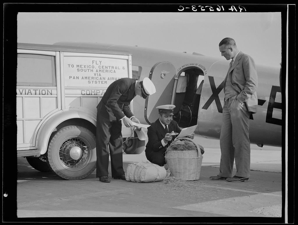 A black and white photo shows three men standing beside an airplane. One man is bending over, appearing to hand a piece of paper or pamphlet to another seated in front of him who reads it as the third looks on with his hands clasped behind his back. Behind them stands a vehicle that says "Fly To Mexico" next to which sits their luggage and baskets filled with what appears to be dried plants.