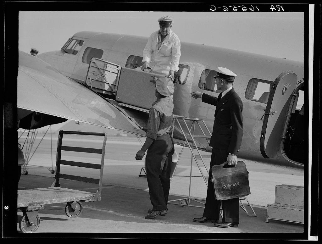 A black and white photograph depicts a scene at an airport during what appears to be the 1930s or early 1940s. There is one man standing on the wing of a propeller-driven aircraft, holding onto its door as if he were assisting someone in boarding or disembarking the plane. He wears light-colored clothing and looks down towards another individual.

On the ground below, two men are engaged with each other; one dressed in dark business attire carrying a briefcase labeled "X-A-BAS", while the other is wearing a pilot's uniform complete with cap and jacket that has insignia or rank markings on it. The man in uniform gestures toward his colleague who holds onto what seems to be an aircraft component, possibly for inspection.

The scene also includes various airport equipment such as wooden pallets known as 'dolly' wheels used for moving cargo around the airplane's wing area, and a metal cart with a flat platform that could have been utilized for carrying luggage or other goods. The backdrop shows more of the aircraft's side profile along with part of its landing gear.

The image has text at the top right corner indicating "D-029451", which seems to be an identification number, and handwritten notes in Arabic script above that might provide additional context but are not legible here.