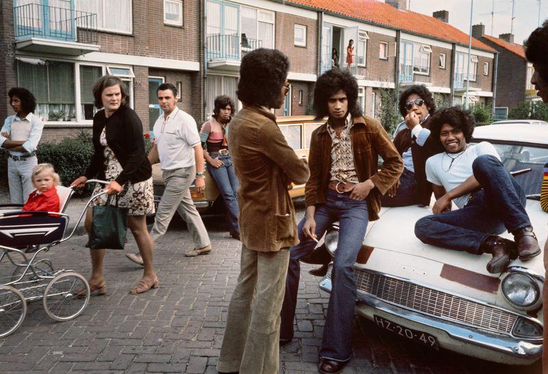 A group of people gathered on a cobblestone street, with the focus on two men sitting casually atop an older model white car. The setting appears to be in a residential area with multi-story brick apartment buildings featuring balconies and windows adorned with shutters. People are engaged in various activities: walking, standing, pushing strollers; some seem engrossed in conversation while others move about the scene unobtrusively. Clothing styles suggest it's likely from an era around the 1970s or '80s, characterized by flared trousers and patterned shirts for both men and women. The mood is relaxed yet vibrant with a mix of interactions reflecting daily life in that period and place.