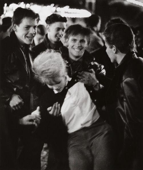 The image is a black and white photograph capturing an outdoor social gathering. It features several young men in casual attire, with one prominently wearing glasses and another laughing heartily. The group appears to be interacting jovially as they embrace or hold onto each other affectionately. In the background, there are blurred lights suggesting this event takes place during nighttime at a lively venue like a market fair or festival. This photograph is by Ed van der Elsken, known for his iconic street photography capturing candid moments of urban life in Amsterdam and beyond.

In terms of accessibility: The image captures an intimate moment among friends, possibly depicting camaraderie and joy found within the bustling environment. Despite being black-and-white, it evokes a sense of connection through shared laughter and physical closeness amidst the lively atmosphere.