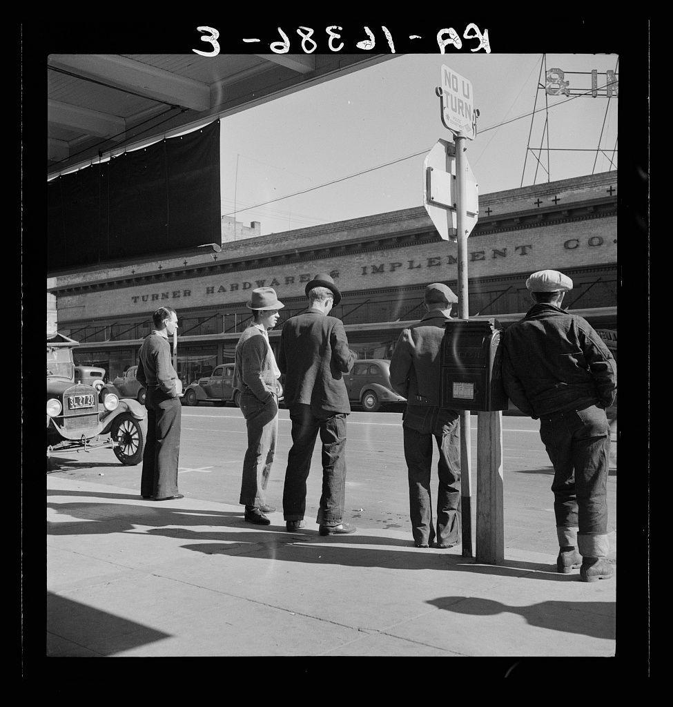 The picture is a black and white photo of four men on the sidewalk in what appears to be an urban area. They are standing near various street signs, including one indicating "No Turn." Behind them, we can see several vintage cars parked along the curb and part of a building with the words "Turner Hardware Implement Co" written across it. The attire suggests this photo is likely from mid-20th century America. Each man has distinct postures: two are leaning against poles or street fixtures, one appears to be engaged in conversation while walking past them, and another stands alone facing away from the group, possibly waiting for someone.

In terms of context provided by "Men on 'Skid Row'." Modesto, California" link - this seems like a social commentary photograph capturing life around what is known as Skid Row. It's an area within large American cities where poverty and homelessness are prevalent issues. The photo could represent the everyday interactions or struggles faced by people in such neighborhoods during that era.

The image captures a slice of urban street life, offering insights into societal conditions through its portrayal of individuals' engagement with their environment.