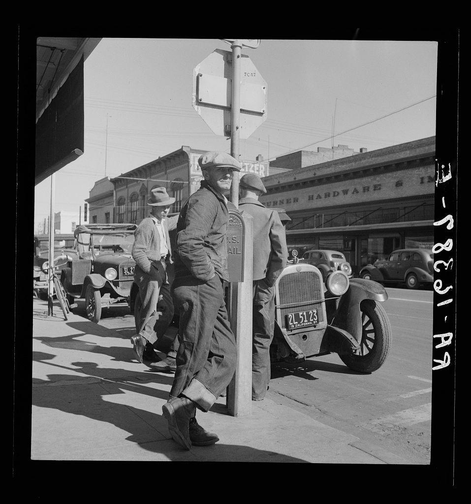 A black and white photograph shows two men on a city street leaning against a pole. The man in the foreground is wearing work clothing including overalls, a hat, shoes with visible laces, and rolled-up pants at the ankles. He has his hands in his pockets while looking directly towards the camera. In the background stands another man also dressed casually but not as prominently featured.

Behind them are parked vintage cars that give an impression of mid-20th century time setting. The street is lined with various businesses, including a hardware store and other establishments indicating it's possibly a commercial area in Modesto, California known as "Skid Row."
