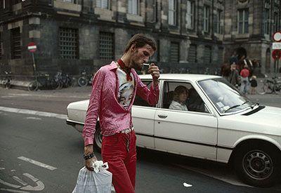 In the image, a young man is seen standing on a street corner. He is dressed in striking red clothing: a long-sleeved shirt and trousers paired with bright pink shoes. The outfit suggests that this might be during the 70s or early 80s when such bold fashion statements were popular among youth.

The man has his right hand raised to his face, holding what appears to be a cigarette in an old-fashioned manner, adding character to his pose and creating a sense of nonchalance. His left arm is bent at the elbow with his fingers extended outwards as if he's about to gesture or point towards something off-camera.

He carries a plastic bag under his right arm which suggests that he may have been shopping recently. In one hand, possibly discarded on the pavement, there seems to be an empty cigarette pack.

Behind him is a vintage white car parked along the curb of the street lined with European-style buildings indicative of urban architecture from earlier decades in Europe or North America. The backdrop also includes pedestrians and bicycles hinting at daily life activities going on around this young individual as he stands out prominently against it all, embodying a youthful spirit amidst everyday city life.

This photo appears to capture not just an instance but a mood - that of rebellion, individualism, and perhaps the fleeting moments of youth. The source for additional informat [...]