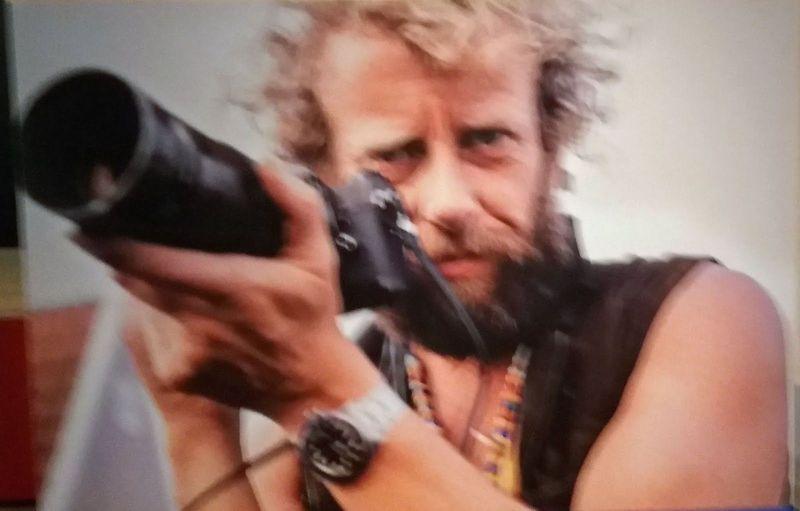 The image depicts a man with curly hair and facial stubble, intensely aiming a large camera lens through his viewfinder. He is wearing sunglasses pushed up onto his head, a black sleeveless vest over what appears to be a patterned shirt, multiple bracelets on one wrist, and has various necklaces around his neck. The background of the image is indistinct but seems to have muted colors with hints of red or pink at the edge, possibly suggesting an indoor setting. Notably, this photograph resembles work by Ed van der Elsken, a Dutch photographer known for capturing candid images from urban environments and documenting social scenes in post-World War II Europe.