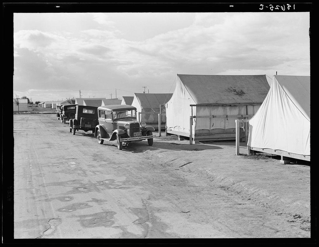 The image depicts a historical scene, likely from the mid-20th century based on the black and white nature of the photograph. A row of vintage automobiles is parked in front of several large tents or temporary structures with canvas walls that appear to be part of a campsite. These could possibly serve as housing for migrant workers at Shafter Camp.
The setting seems arid, indicated by the sandy ground and overcast sky above. The vehicles are old-fashioned models from an earlier era, suggesting this photograph captures life during a time when such automobiles were common on roads. This scene might be part of a larger collection documenting living conditions in camps meant for migrant workers engaged in agriculture or related labor.
The tents serve as makeshift accommodations while the occupants supply their own personal items and belongings. Some newer camp designs included prefabricated steel one-room houses, aiming to provide better living arrangements compared to canvas structures like those pictured here.