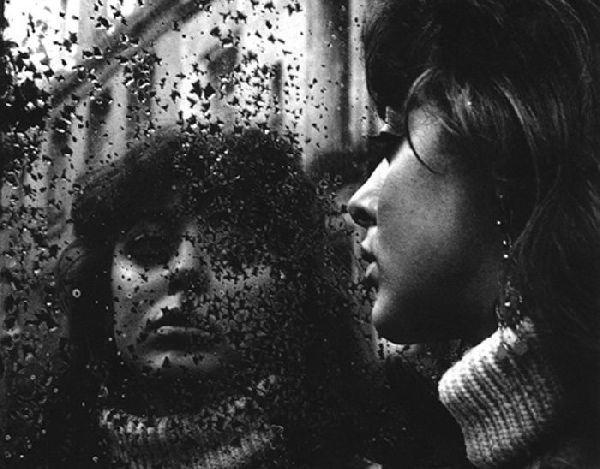 The image is a black and white photograph featuring two individuals with their faces partly obscured by what appears to be raindrops on the window they are looking through. The person in the foreground has curly hair, wears large earrings, and is dressed in a turtleneck sweater or scarf. Their gaze seems contemplative as it looks outwards.

The second individual's face is partially visible behind the first one; this person also appears to be wearing eyeglasses with a reflective surface that adds depth to their image. The overall mood of the photograph evokes introspection and perhaps melancholy, given the weather conditions implied by the raindrops on the glass.

Additional information about this image can be found in "Ed van der Elsken | Donkere fotografie" (Dark Photography) under EdvdElsken at the provided URL.