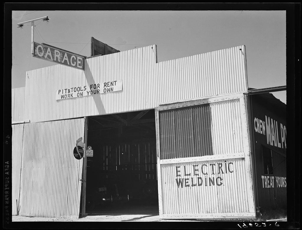 The image is a black-and-white photograph depicting an industrial or workshop setting, specifically highlighting portable metal structures used as garages and storage spaces. The dominant structure reads "CARAGE" at the top with additional text stating that pit and tools for rent are available on site, encouraging individuals to work on their own projects. Another sign indicates availability of electric welding services. Additional smaller signs indicate specific areas such as "CHEMMAIL PO," though it's unclear what this stands for within this context.

The structure appears weathered and utilitarian, with a simple metal construction that suggests functionality over aesthetic appeal. The photograph is credited to Dorothea Lange, known for her work during the Dust Bowl era in America, capturing subjects of social documentary photography such as migrant workers, rural poverty, homelessness, droughts, industrial laborers, African Americans, and other minorities.

The timestamp "1932" suggests it was taken more than a century ago. The photograph captures an aspect of American industry or resourcefulness during that period, likely in California given the reference to Fresno County. This image is part of Lange's extensive body of work documenting various aspects of life across America during the early 20th century.

The source link indicates it might be related to a collection  [...]