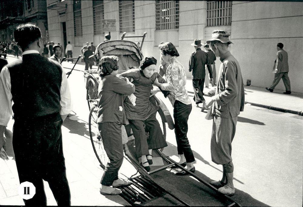 The image captures a lively street scene in Hong Kong during the late 1950s, as suggested by its historical context. It is presented in black and white, adding to the vintage feel of the photograph.

In the foreground, two women are engaged in conversation; one seems to be comforting or consoling the other with her arm around her shoulders. A young girl accompanies them on a rickshaw pulled by an individual dressed in traditional attire, indicating the integration of local customs and urban life.

On the right side, another woman stands beside someone wearing casual clothing and carrying what appears to be newspapers under their arm, perhaps selling or distributing newsprint from that era.

The background features various pedestrians moving along the sidewalk, some walking alone while others are engaged in conversations. The architectural structures lining the street suggest a blend of old and new with barred windows on one building hinting at historical security measures.

A man wearing glasses is seen standing to the left side of the frame, observing or possibly waiting for someone. His presence adds another layer to the dynamic atmosphere captured within this snapshot of Hong Kong's urban life from that period.

The image serves as a visual narrative encapsulating daily interactions and activities in late 1950s Hong Kong, showcasing both individual stories and collecti [...]