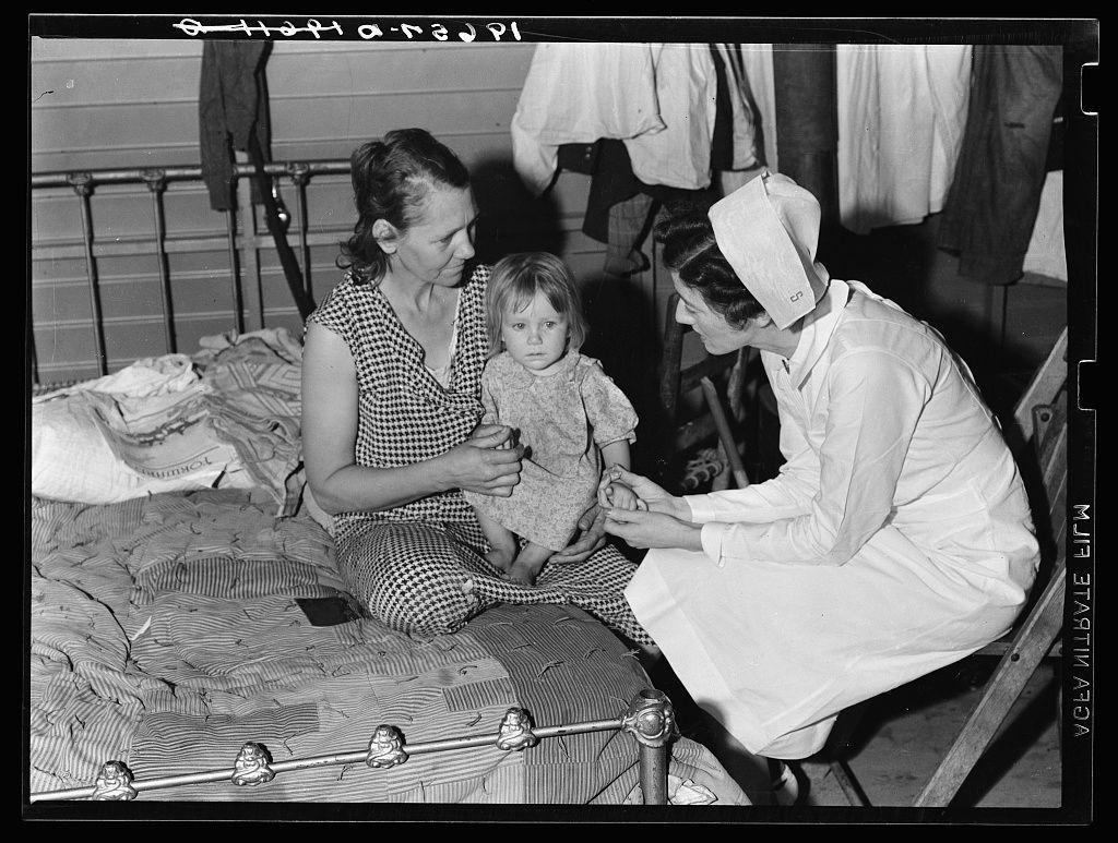 The image depicts a historical scene with three individuals in an indoor setting, likely from the early to mid-20th century. On the left, there is a woman seated on what appears to be a bed or cot. She has her hair pulled back and wears checkered pajamas over dark clothing. In her lap sits a young child dressed in patterned onesie with puffed sleeves.

In front of them, kneeling beside the child's leg, is another woman wearing nurse attire: white apron, cap, and coat. Her attention seems focused on assisting or examining the baby. The setting suggests a domestic environment, possibly within an institutionalized healthcare context given her professional dress code.

The backdrop includes what looks like hanging laundry items above them, indicating personal living quarters in close proximity to medical care facilities. The lighting is natural but not overly bright, suggesting this photograph was taken during daylight hours without direct sunlight flooding the room.