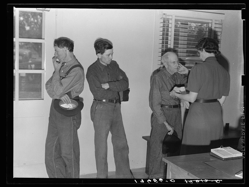 The image depicts four individuals in what appears to be an indoor setting with a window and blinds visible on the right side. Two men are standing against the wall, one of whom is holding his hat while conversing over the phone, identifiable by the receiver held close to his ear. The other man stands beside him with arms crossed, exhibiting a contemplative or possibly anxious posture.

To their left, another young man looks directly at the camera, seemingly engaged in conversation or waiting for something. Across from them on the right side of the frame is an older couple; the woman wears glasses and appears to be speaking to the seated elderly man, who has his eyes closed as if resting or listening intently.

A table with a few items such as papers or possibly medical supplies can be seen in front of this group. The image's black-and-white nature suggests it could be historical documentation from an earlier era.