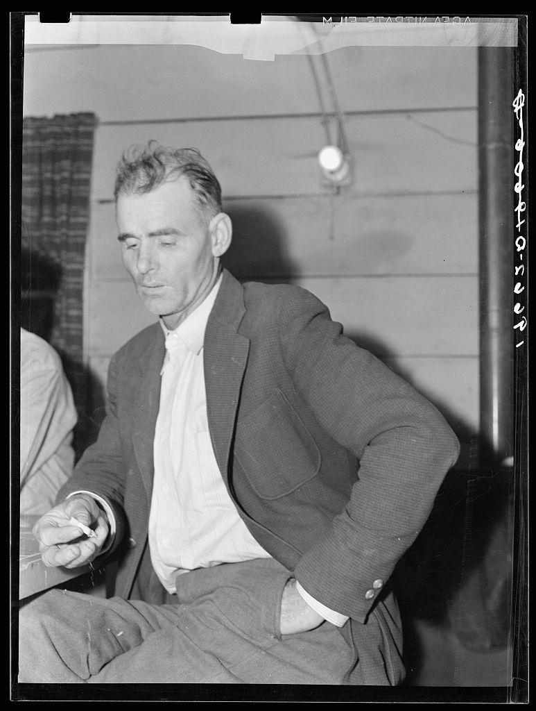 The image is a black and white photograph depicting an older man seated indoors. He appears contemplative, with his head slightly bowed down as he gazes off to the side, not directly engaging with the camera or viewer. The man has tousled gray hair, prominent on the top of his head but receding at the temples.

He is dressed in a casual yet neat manner: wearing a dark blazer over a light-colored shirt without a tie, and rolled-up trousers that reveal a bit more leg than usual for formal wear. His right hand holds an object which seems to be a pen or small cigar, suggesting he may have been writing or smoking before the picture was taken.

The background is minimally detailed but reveals part of an interior setting with what appears to be curtains and possibly some other furnishings that are not clearly discernible due to focus on the man. The lighting casts shadows across his face and jacket, creating a contrast between light and shadow which adds depth to the image's composition.