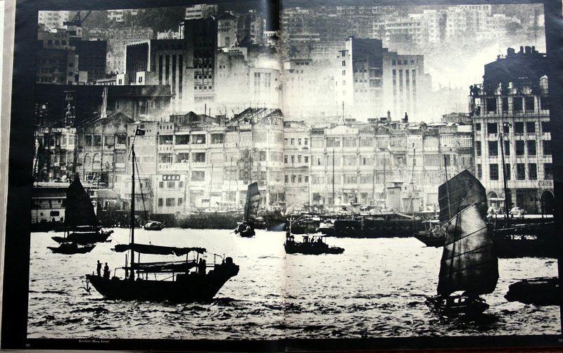 The image is a black and white photograph depicting an urban harbor scene. It shows several sailboats with large, billowing sails in the foreground on choppy water, suggesting movement or possibly wind. These boats are likely traditional junk vessels commonly used for transportation within Hong Kong's waters.

In the background, there's a dense arrangement of mid-20th-century buildings varying in architectural styles and states of repair. Some structures appear dilapidated with visible damage, hinting at historical wear or possible wartime effects. The skyline is interrupted by tall modern skyscrapers that stand out against the older architecture, indicating urban development over time.

The composition has a sense of depth, with boats closer to the viewer on the lower right and buildings receding into the distance towards the upper left corner. This juxtaposition suggests a bustling port area transitioning from historical elements to contemporary growth.

There is no visible text or discernible branding within this photograph that would suggest it was taken by any specific photographer aside from Ed van der Elsken, as per the provided additional information indicating its connection with him and Hong Kong Harbor in 1960. The quality of the image suggests a vintage source, consistent with what might be expected for an early to mid-20th-century documentary photograph captu [...]