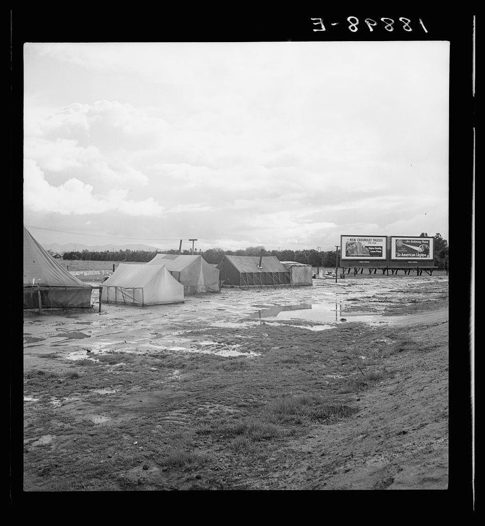 The image is a black and white photograph depicting an outdoor scene with several temporary structures, including tents and prefabricated buildings. The ground appears to be muddy or wet in places, suggesting recent rain or poor drainage conditions.

In the background, there are billboards displaying advertisements for products such as "Fruitland" cigarettes and other unspecified items. The sky is partly cloudy, indicating a potentially overcast day. There's an overall impression of a makeshift campsite, possibly used by workers during off-seasons or temporary work situations in agricultural areas.

Additional information about the image indicates that it was taken near Lindsay, California, specifically among citrus workers who were occupying winter quarters in Tulare County. The reference to Dorothea Lange suggests this photo is part of her extensive body of work documenting various aspects of American life during and after World War II through the Farm Security Administration (FSA) program.

The photograph captures a moment that reflects on labor conditions, living arrangements for migrant workers, and perhaps broader themes related to agricultural cycles and seasonal worker housing.