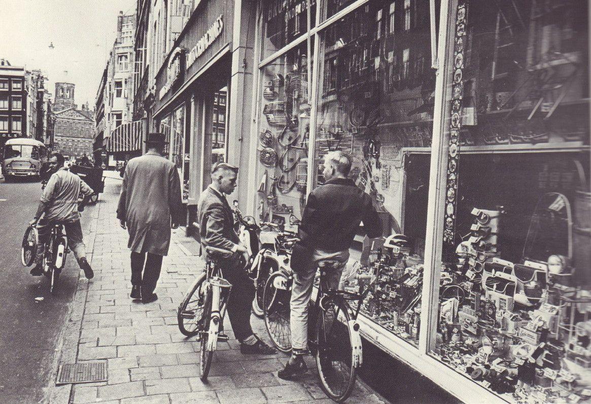 The image is a black and white photograph depicting an urban street scene from what appears to be the late 20th century. Several individuals, including men in jackets and hats, are seen on bicycles along a pavement lined with shops. The storefronts display various items such as cameras, glasses, and other goods for sale. A few pedestrians walk by or stop near the shop windows, seemingly browsing through the displayed products. The architecture of the buildings suggests an older European cityscape, potentially from post-war years considering their style and condition.

The photo captures a moment in everyday life with people engaging in leisurely activities like cycling and window shopping. This scene might reflect a typical day in Amsterdam during that time period, as indicated by the title "Amsterdam! Oude Foto's -1947-1970" (Amsterdam: Old Photos - 1947-1970) mentioned in the source information provided.

The image has historical significance and offers a glimpse into urban life during that era, showcasing social interactions, transportation methods such as bicycles, and commercial activities within a European city.