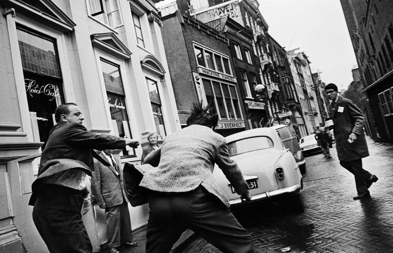 The image is a black and white photograph capturing an urban street scene. In the foreground, two men appear to be in motion; one man seems to have just thrown something at another person who's turning away from him with their back towards us. The first man wears dark clothing while the second sports a lighter-toned jacket over a darker shirt.

In the middle of the image is an older model white car parked on the street, which has been labeled as "31" indicating it might be part of some system or event. A third person is seen in profile walking away from the action towards us and appears to be wearing glasses with dark clothing.

The background shows a row of classical European-style buildings with storefronts like 'Hotel City' on the left, suggesting this scene takes place in an urban environment possibly within Amsterdam given the context provided by the URL. The architecture indicates historic streets that could belong to cities such as those found in Western Europe during mid-century times. There's also another individual walking down the street wearing a coat and hat.

The photo gives off a candid feel, capturing spontaneous interactions among people on a city sidewalk. It seems like an everyday scene with no specific focal point other than the action between two individuals near the parked car.