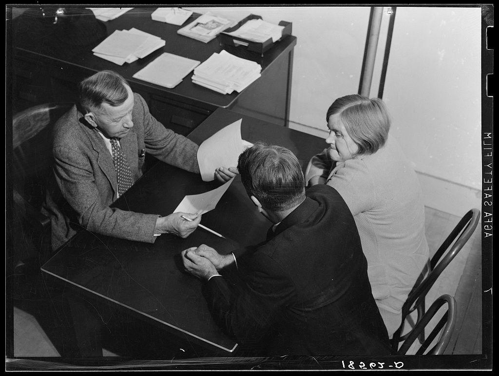 The image captures a moment in an office where three individuals are seated around a table. The scene appears to be from the mid-20th century, as suggested by their attire and the black-and-white photographic quality of the photo.

On the left side of the frame is a man wearing a suit with a patterned tie, deeply engrossed in examining some papers held within his hands. His expression suggests concentration or analysis. Opposite him sits another individual dressed in dark clothing, also focused on perusing documents while holding a pen ready to write or mark something.

To their right stands an older woman observing the interaction between them with interest. She appears contemplative and engaged, possibly waiting for her turn to speak or contemplating what she has heard so far.

The table is cluttered with stacks of papers, indicating active work being conducted here. A chair can be seen on the left side of the frame, which might belong to another person who was part of this meeting but not currently visible in the image.

In summary, the photograph documents a serious discussion among three individuals likely involved in some official business or decision-making process within an office setting from the past century.